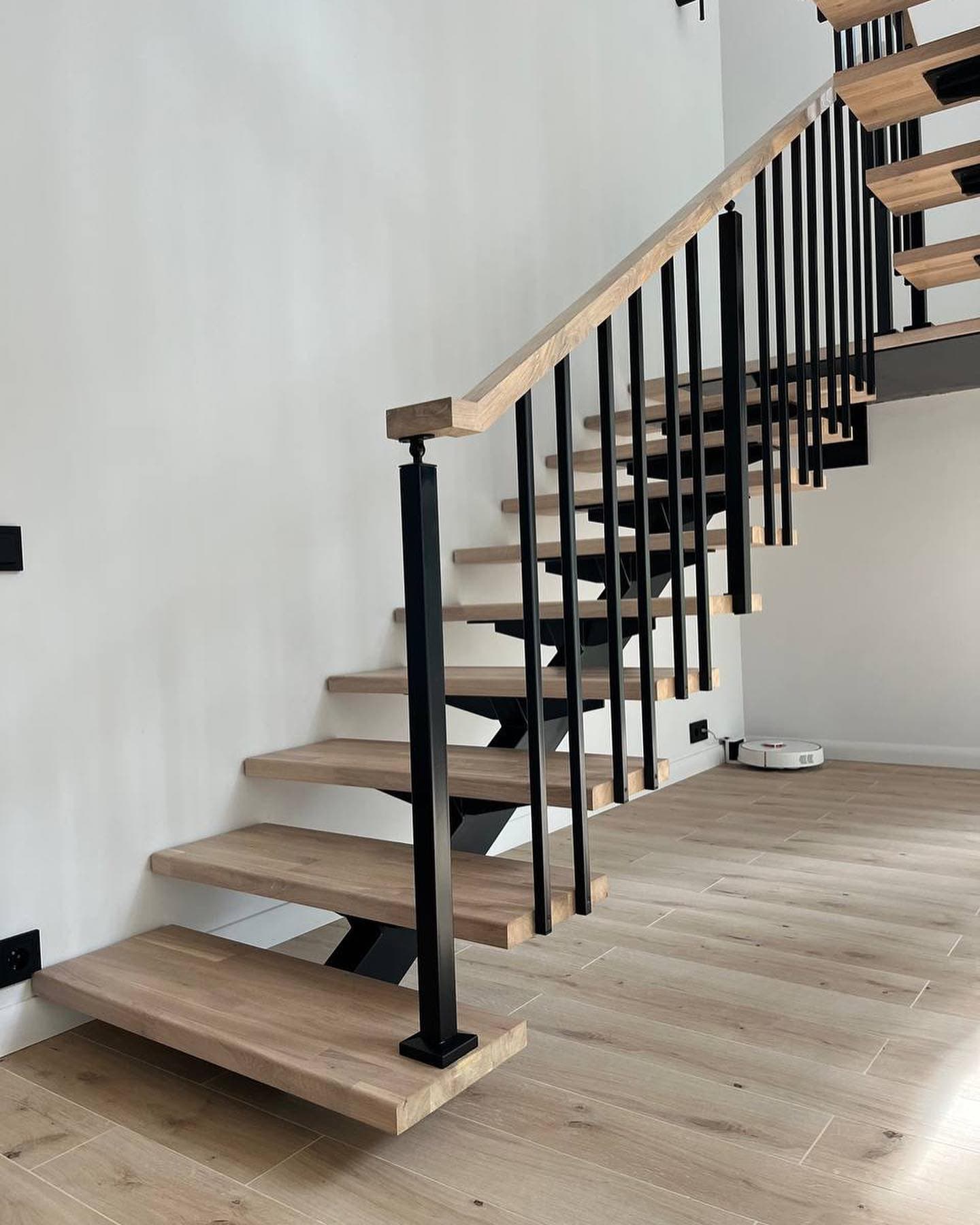Modern floating staircase with light wood treads and black metal railing against a white wall.