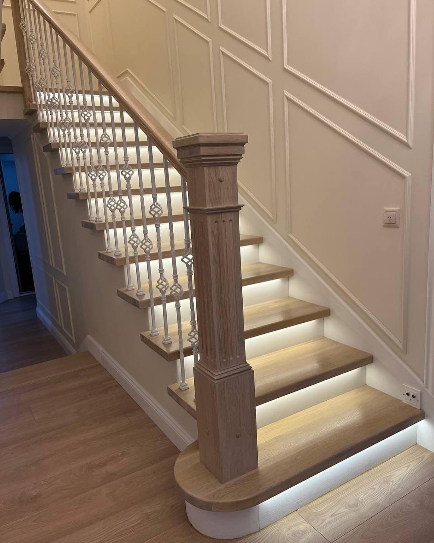 Modern wooden staircase with integrated LED step lighting, white decorative balusters, and wall paneling.